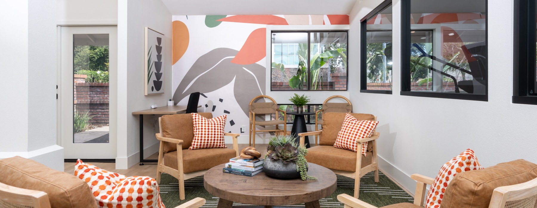Modern lounge with tan armchairs, orange accent pillows, a round wood coffee table, and a colorful abstract mural.