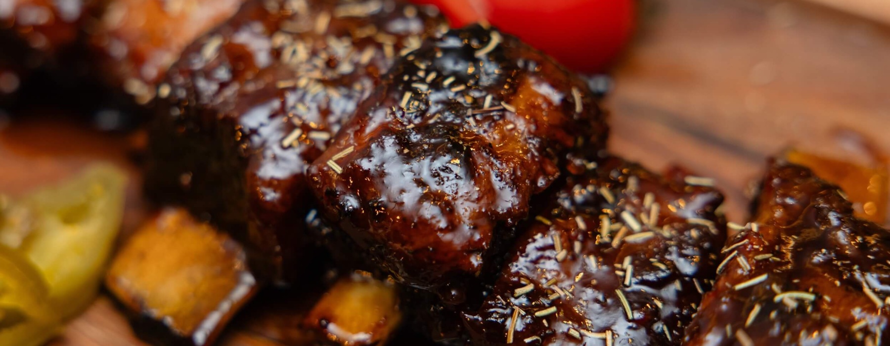 Close-up of glossy barbecue-glazed rib pieces sprinkled with dried herbs, served on a wooden board with a whole tomato and pickle slices in the background.