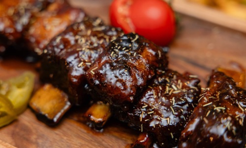 Close-up of glossy barbecue-glazed rib pieces sprinkled with dried herbs, served on a wooden board with a whole tomato and pickle slices in the background.