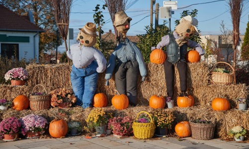 Three scarecrows among hay bales, pumpkins, mums, and baskets in an outdoor fall display.